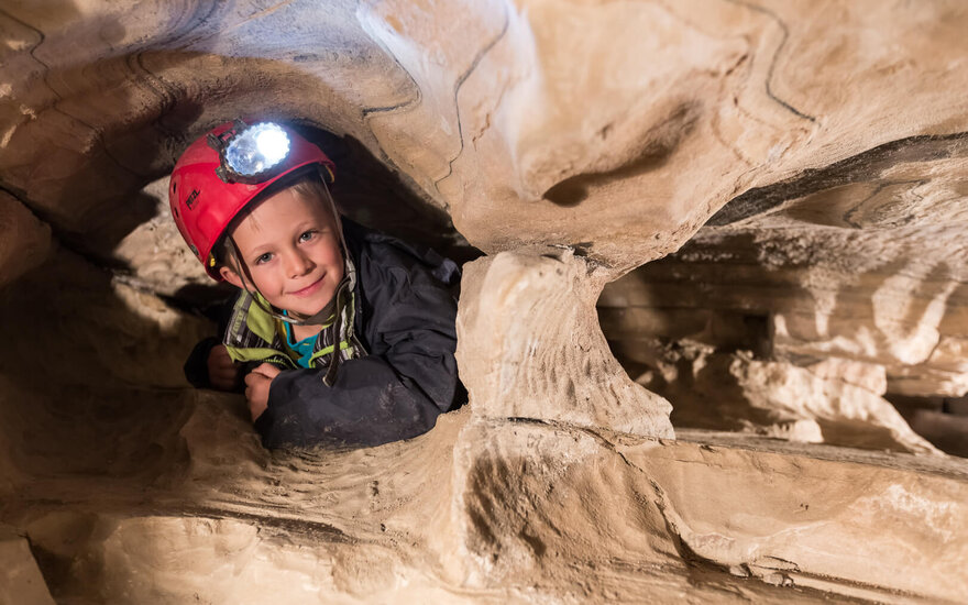 Ein Junge mit einem roten Helm lächelt, während er in einer engen Höhle sitzt und von einer Lampe beleuchtet wird.