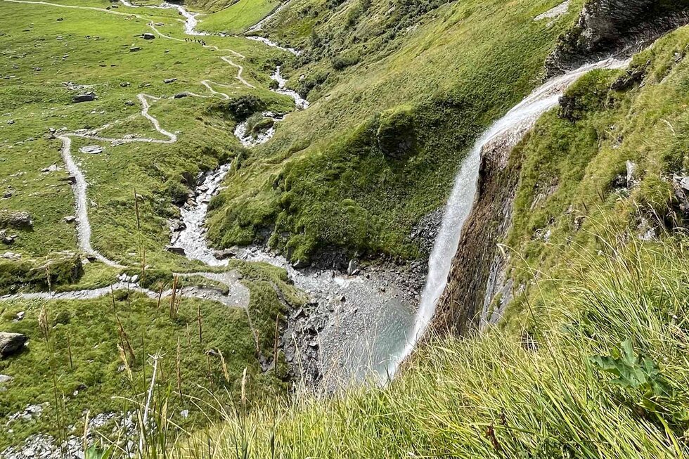 Ein Wasserfall stürzt über eine grüne Klippe, umgeben von saftigem Gras und einem klaren, plätschernden Bach.