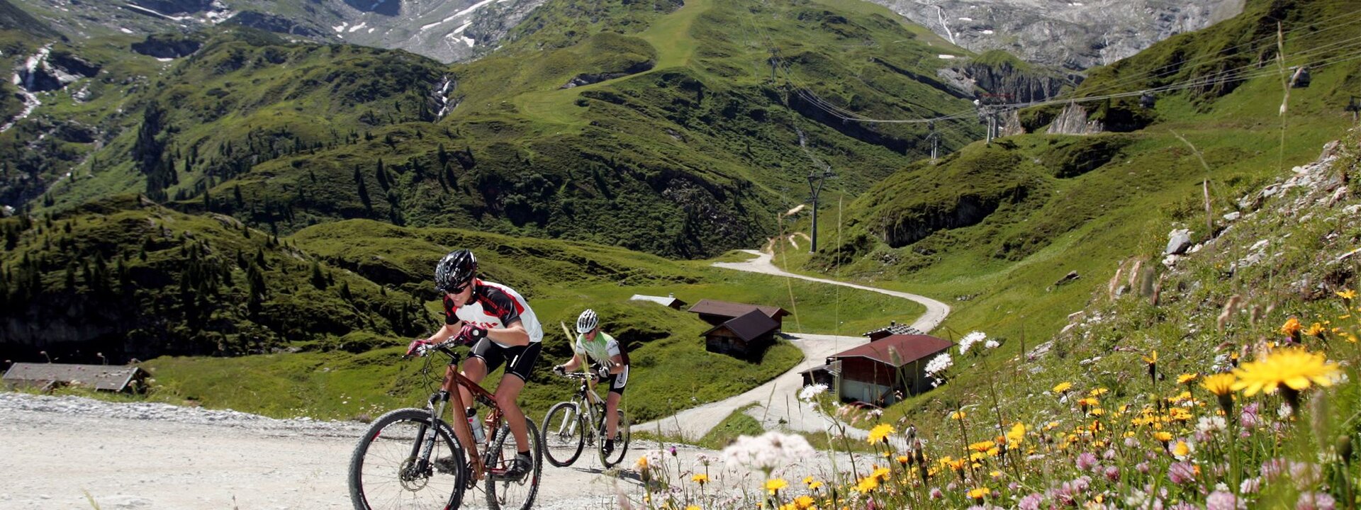 Die Radfahrer kämpfen sich den Hang hinauf, umgeben von saftigem Grün und bunten Blumen, mit Hütten im Hintergrund.