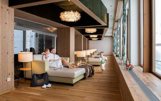 Ein Paar in Bademänteln entspannt auf einer Couch, während ein Kind fröhlich im Spa-Bereich herumläuft.