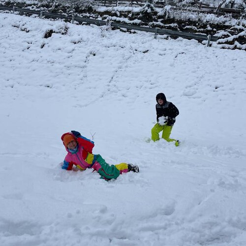 Zwei Kinder spielen im frischen Schnee, eines liegt lachend im Schnee, während das andere aufrecht steht und einen Schneeball hält.