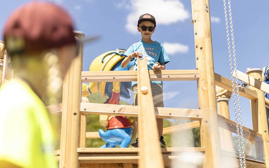 Ein Junge mit Sonnenbrille steht auf einem Spielturm und hält einen Ball in der Hand, während er lächelt.
