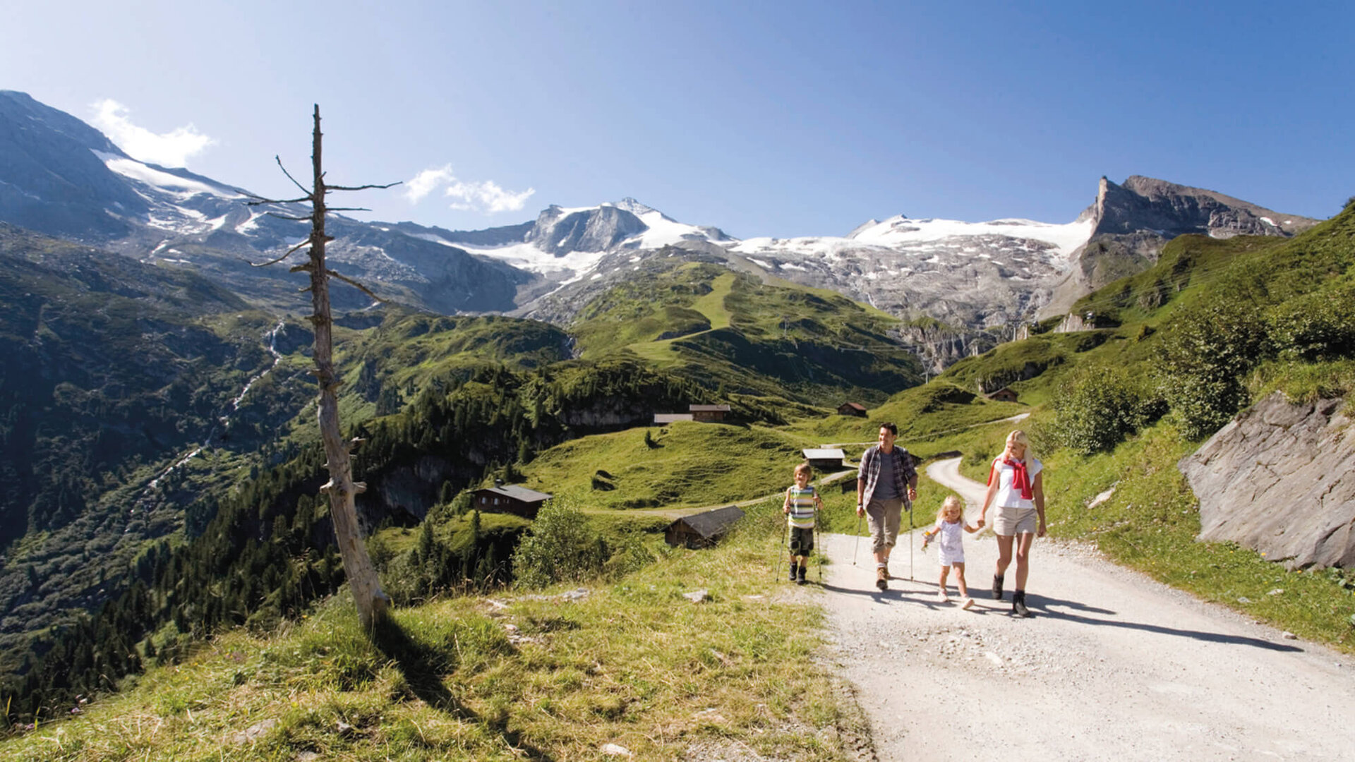 Eine Familie wandert auf einem malerischen Bergweg, umgeben von grünen Wiesen und schneebedeckten Gipfeln.