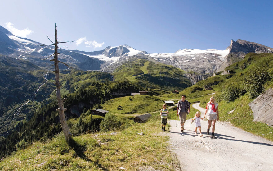 Eine Familie wandert auf einem malerischen Bergweg, umgeben von grünen Wiesen und schneebedeckten Gipfeln.