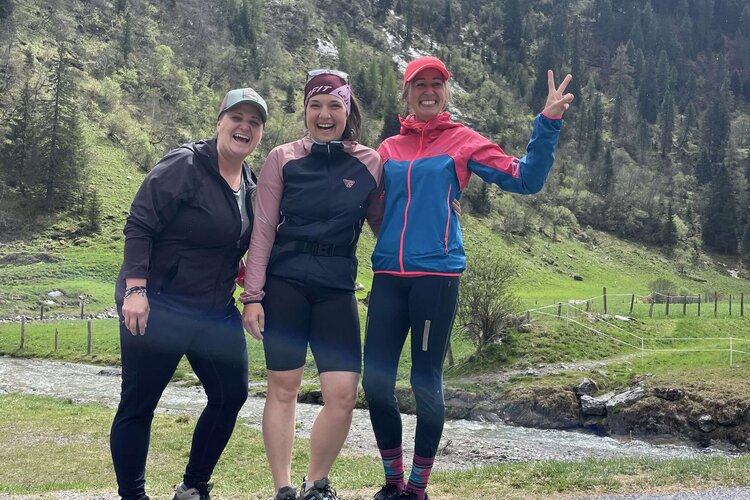 Drei Frauen stehen fröhlich auf einem Weg, umgeben von grünen Bergen und Bäumen, während sie in die Kamera lächeln.