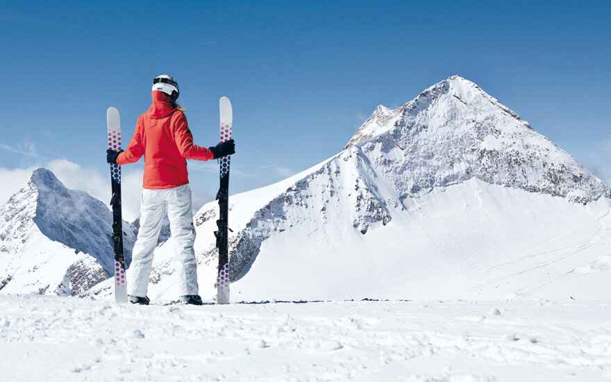 Eine Person in einem roten Anorak steht auf einem schneebedeckten Berg und hält Skier in den Händen, während sie die Aussicht genießt.