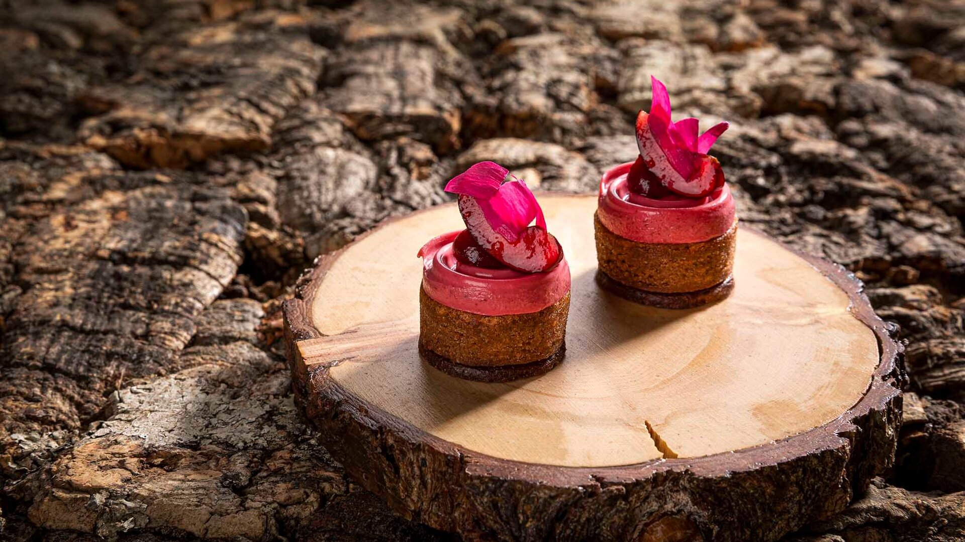 Zwei kunstvoll dekorierte Mini-Törtchen auf einem Holzbrett, umgeben von einer rustikalen Baumrinde.