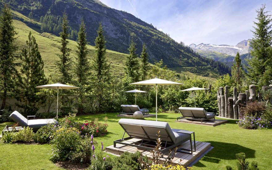 Eine ruhige Gartenlandschaft mit Liegen und Sonnenschirmen, umgeben von üppigem Grün und majestätischen Bergen im Hintergrund.