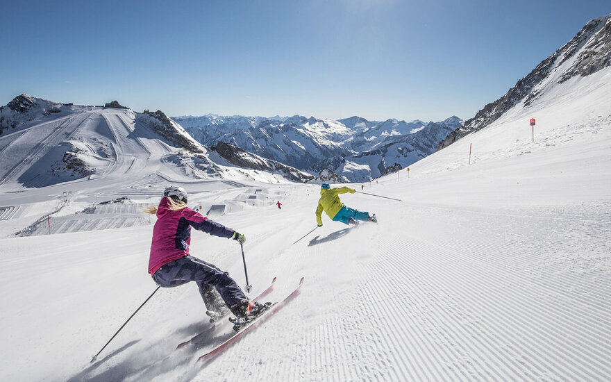 Zwei Skifahrer, einer in einer pinken Jacke und der andere in einer gelben Jacke, fahren auf einer schneebedeckten Piste.