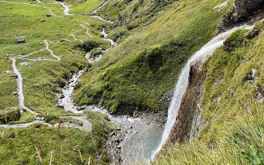 Ein malerischer Wasserfall fließt über grüne Hügel, umgeben von sanften Wegen und glitzerndem Wasser im Tal.
