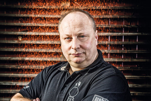 Ein Mann mit ernstem Gesichtsausdruck steht vor einer rustikalen Wand, während Gewürze in der Luft schweben.