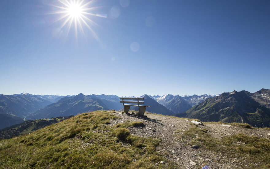 Eine einsame Bank steht auf einem Hügel mit atemberaubendem Blick auf die umliegenden Berge und den strahlend blauen Himmel.