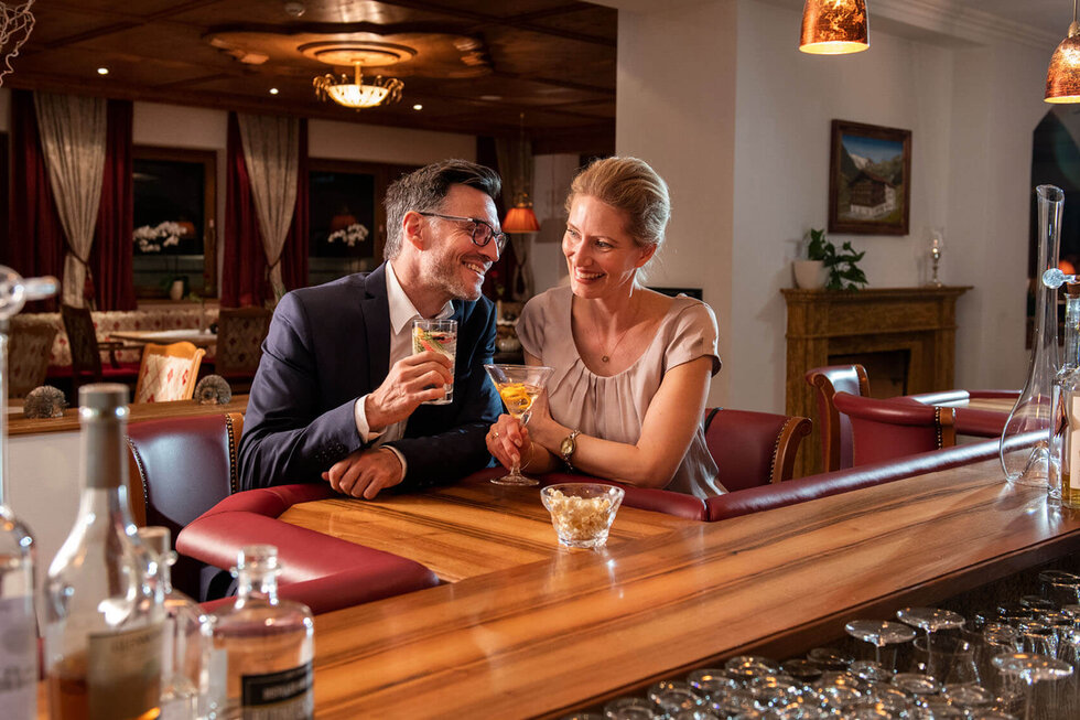 Ein Paar genießt Cocktails an der Bar, umgeben von elegantem Interieur und warmem Licht, während sie sich anlächeln.