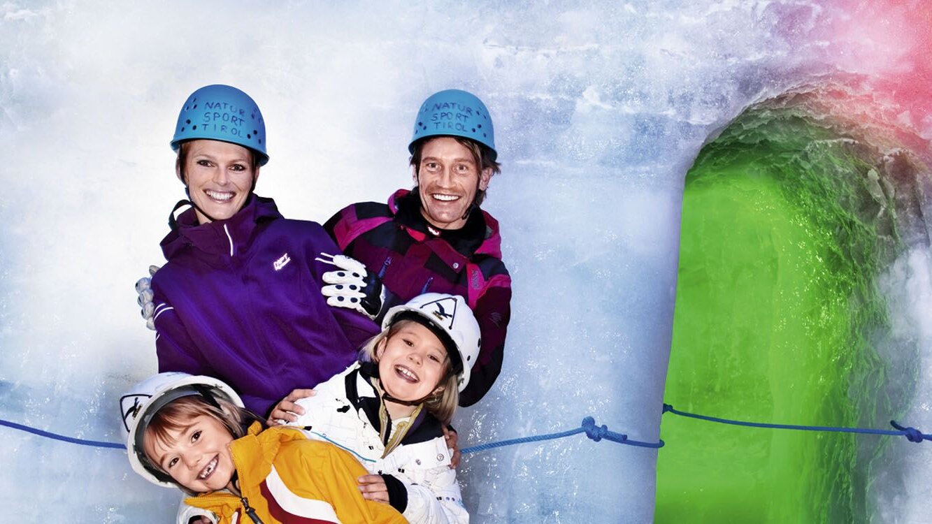 Vier Personen, darunter zwei Kinder, lächeln in einem Eisraum, umgeben von bunten Eisstrukturen und blauen Seilen.