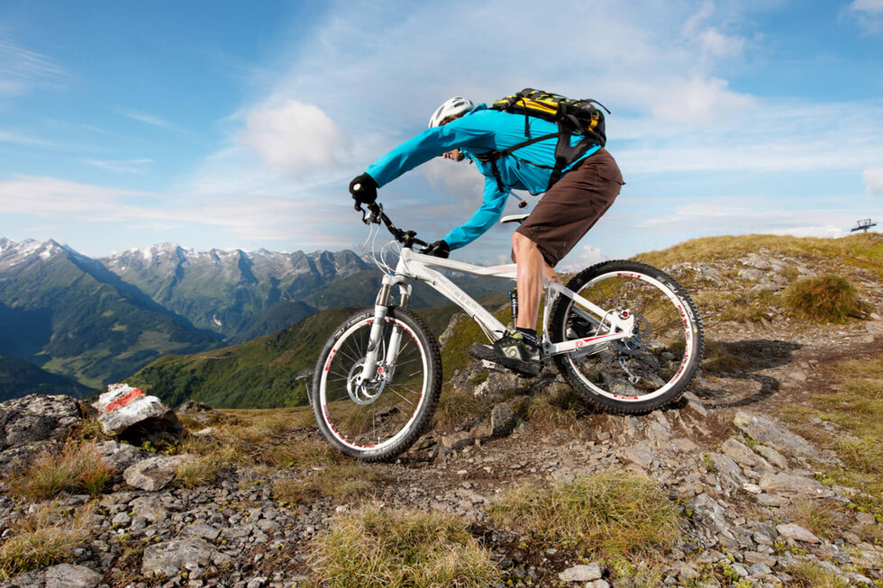 Ein Mountainbiker in einer türkisfarbenen Jacke fährt über einen steinigen Pfad mit beeindruckender Berglandschaft im Hintergrund.