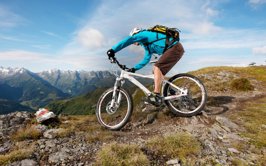 Ein Mountainbiker in einer türkisfarbenen Jacke fährt über einen steinigen Pfad mit beeindruckender Berglandschaft im Hintergrund.