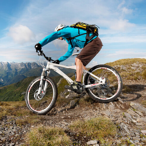 Ein Mountainbiker in einer türkisfarbenen Jacke fährt über einen steinigen Pfad mit beeindruckender Berglandschaft im Hintergrund.