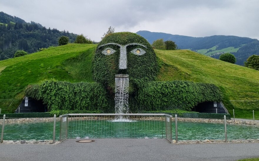 Inmitten einer malerischen Landschaft erhebt sich eine grüne Figur mit Wasserfall, umgeben von sanften Hügeln und Bäumen.