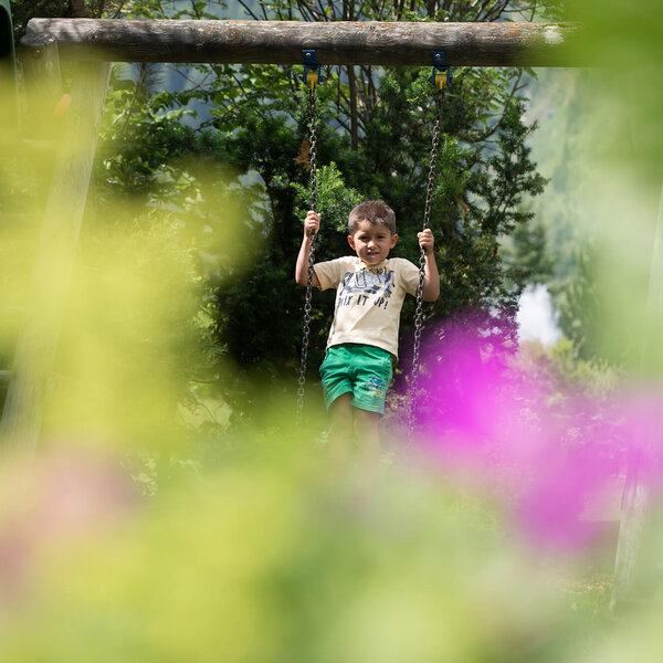Ein fröhlicher Junge schwingt auf einer Schaukel, umgeben von bunten Blumen und grünem Laub im Hintergrund.