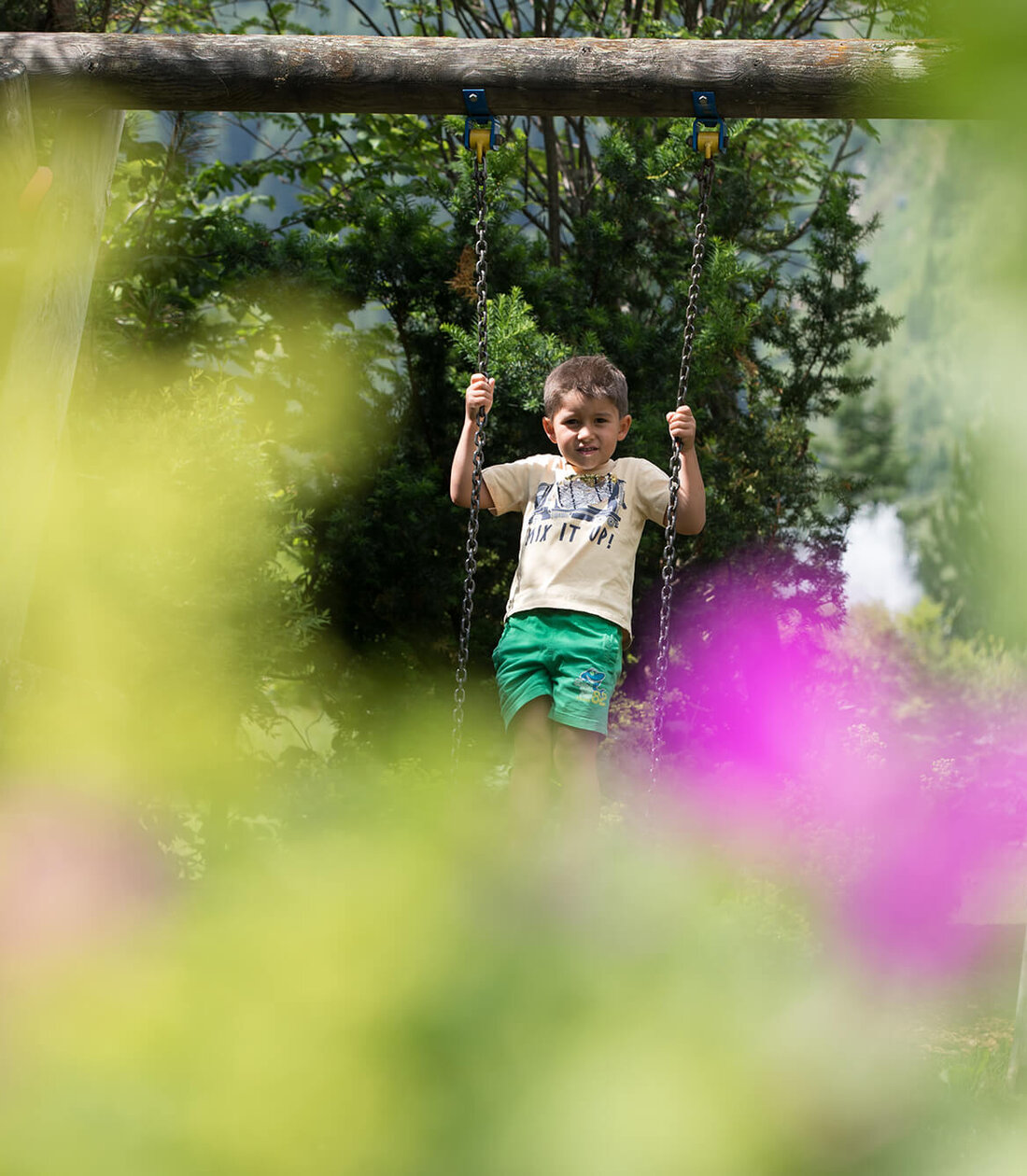 Ein fröhlicher Junge schwingt auf einer Schaukel, umgeben von bunten Blumen und grünem Laub im Hintergrund.