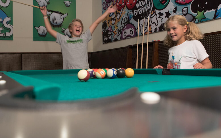 Die Kinder genießen eine Billardpartie, der Junge strahlt vor Freude, während das Mädchen aufmerksam zuschaut.