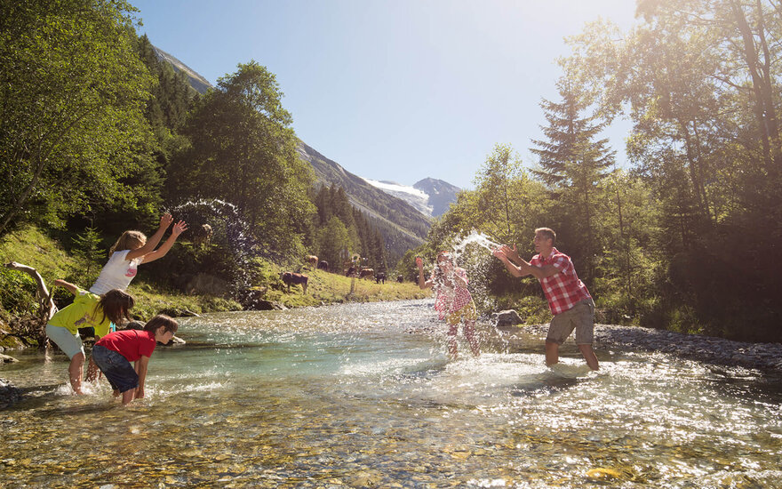 Eine fröhliche Familie spielt im klaren Wasser eines Flusses, während die Sonne strahlt und die Berge im Hintergrund sichtbar sind.