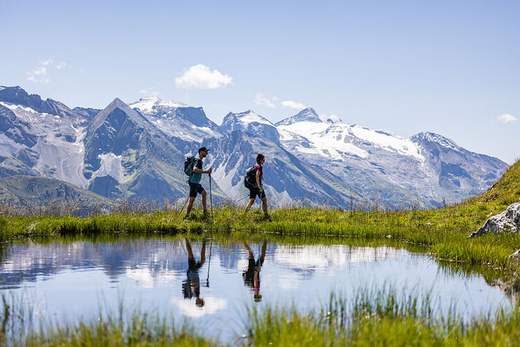 In der alpinen Landschaft wandern zwei Personen, während die schneebedeckten Gipfel im Hintergrund strahlen.