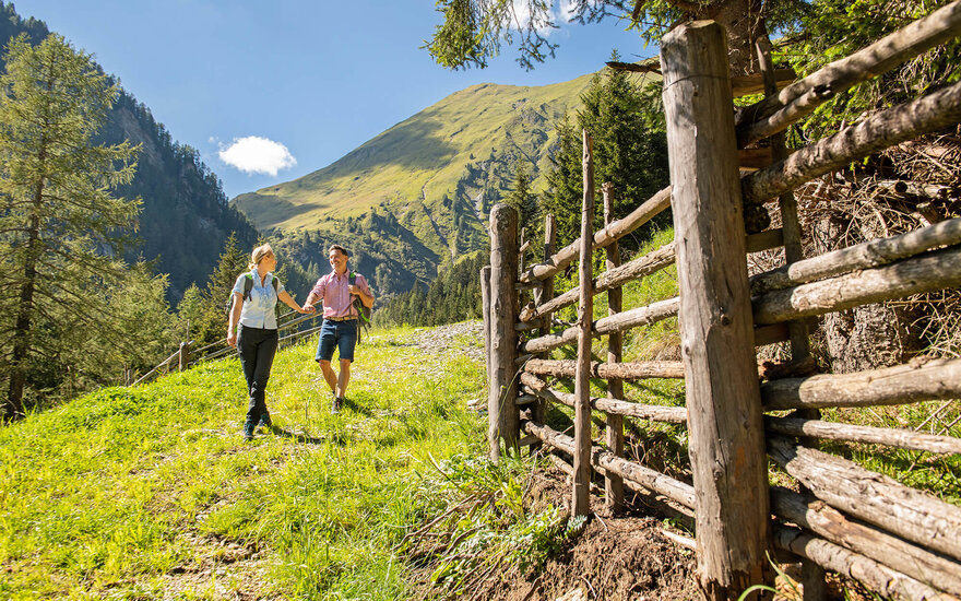 Ein Paar wandert fröhlich auf einem grünen Pfad, umgeben von Bergen und Bäumen, während sie sich an den Händen halten.