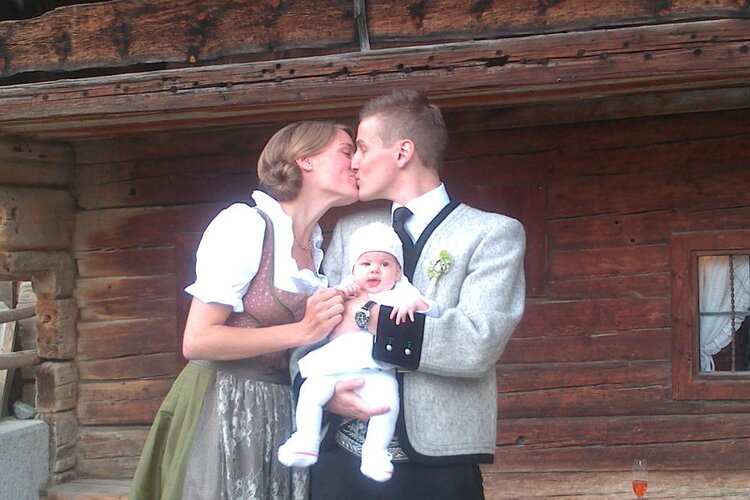 Ein Paar küsst sich liebevoll, während sie ihr Baby in den Armen halten, vor einer rustikalen Holzhütte mit blühenden roten Blumen.