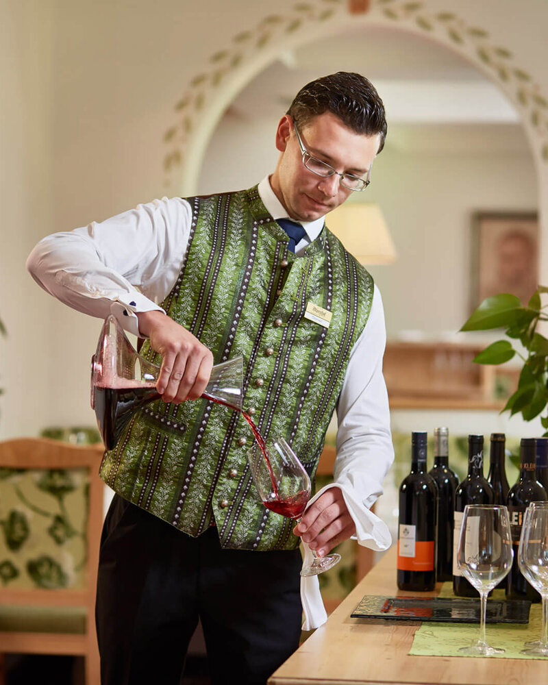 Ein Kellner in einem eleganten grünen Weste schenkt Rotwein in ein Glas, während er in einem stilvollen Restaurant arbeitet.