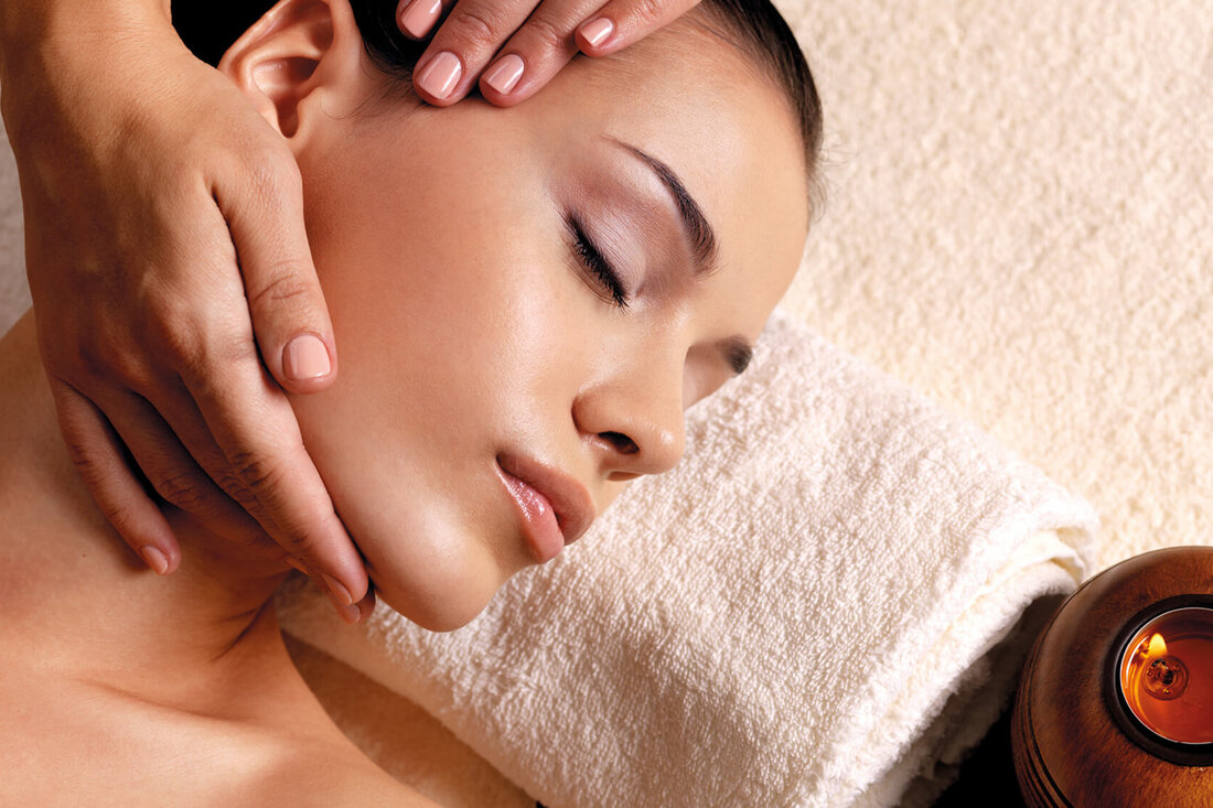 Eine entspannte Frau erhält eine Gesichtsmassage, während ihre Augen sanft geschlossen sind und eine Kerze im Hintergrund brennt.