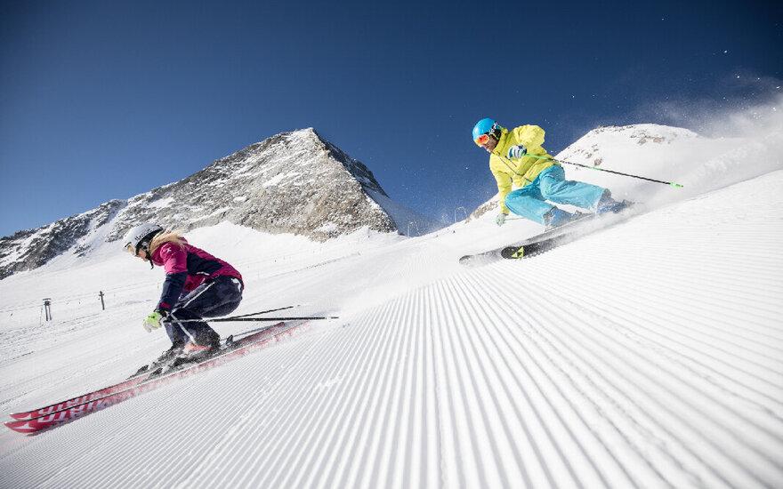 Zwei Skifahrer rasen auf einer perfekt präparierten Piste in den Alpen, umgeben von majestätischen Bergen und klarem Himmel.