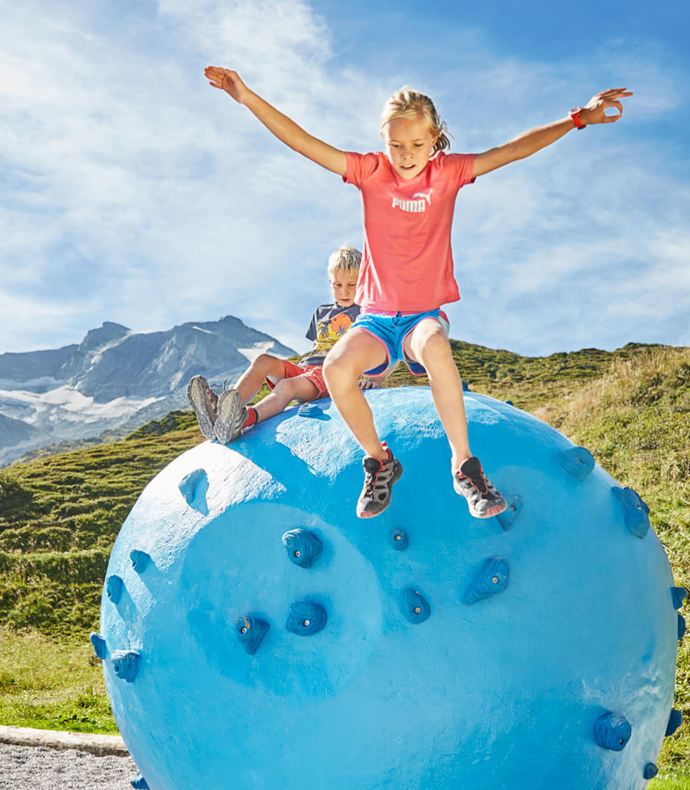 Ein Mädchen springt fröhlich von einem großen blauen Kletterball, während ein Junge im Hintergrund sitzt und zuschaut.