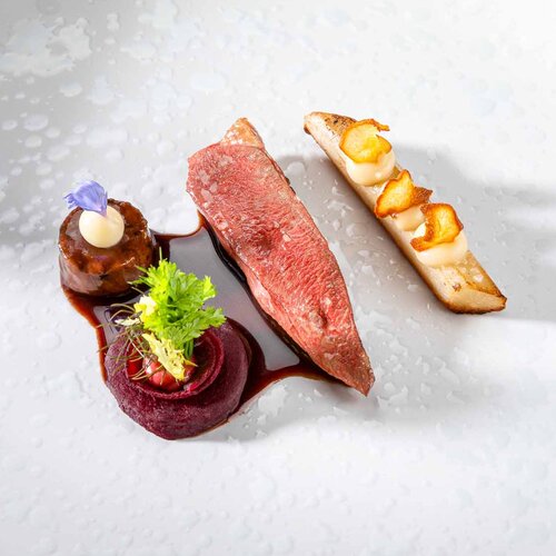 Ein kunstvoll angerichteter Teller mit zartem Fleisch, Gemüse und einer Sauce, umgeben von dekorativen Elementen.