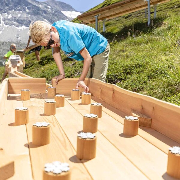Ein Junge mit blonden Haaren und Sonnenbrille spielt an einem Holzspielgerät in den Bergen, umgeben von grüner Wiese.