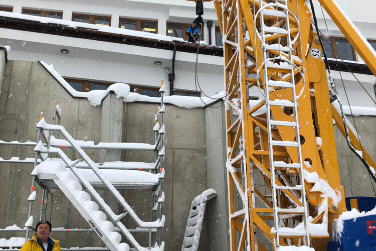 Ein Bauarbeiter steht vor einem Kran, umgeben von Schnee, während er die Arbeiten an einem Gebäude überwacht.
