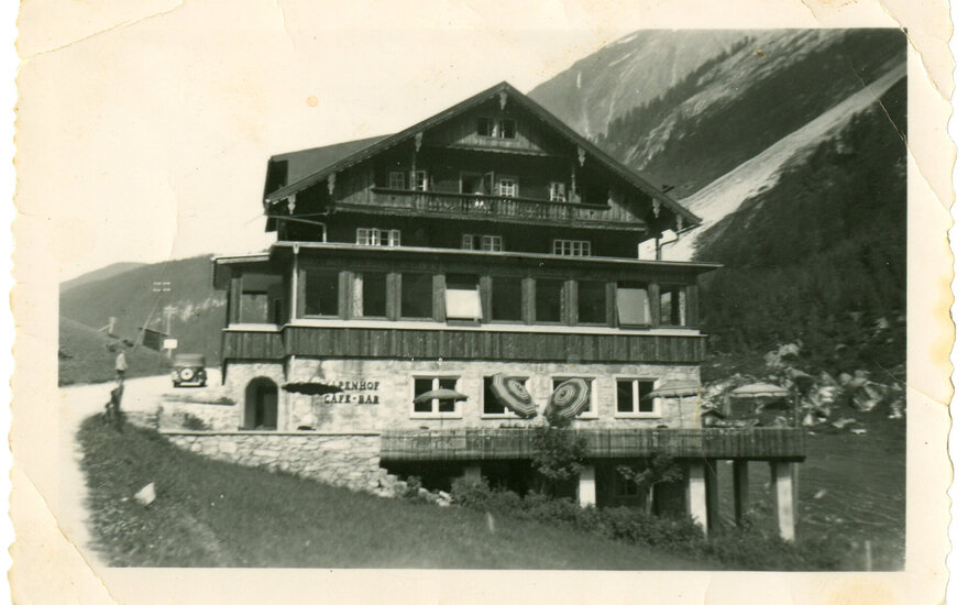 Ein traditionelles Berghaus mit Holzverkleidung, umgeben von grünen Wiesen und majestätischen Bergen im Hintergrund.