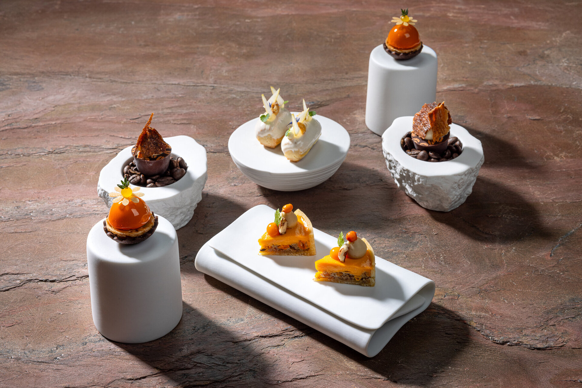 Elegante Desserts auf weißen Tellern, darunter ein schokoladiges Gebäck und fruchtige Törtchen, umgeben von Kaffeebohnen.