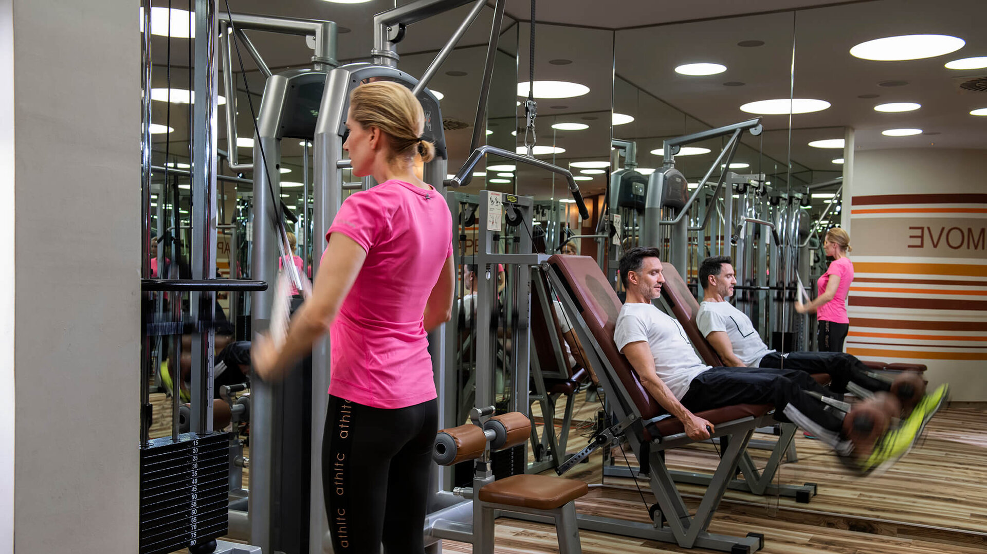 Eine Frau in einem Fitnessstudio trainiert an einem Kraftgerät, während zwei Männer auf einer anderen Maschine sitzen und sich unterhalten.