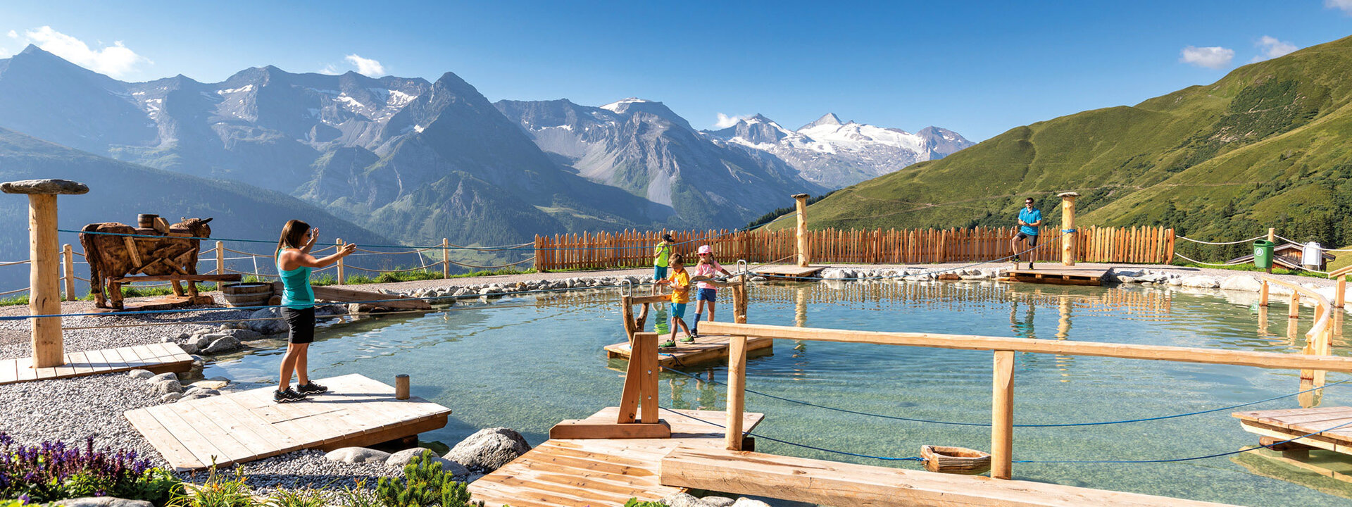 Eine Frau steht am Ufer eines klaren Gewässers und macht ein Foto, während Kinder auf Holzplattformen spielen.