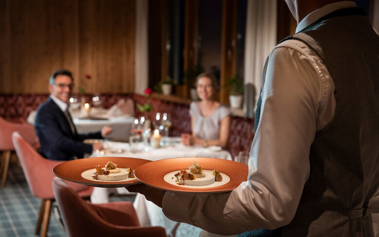 Ein Kellner serviert elegant zwei Teller mit feinem Essen an einem Tisch, während ein Paar im Hintergrund lächelt.
