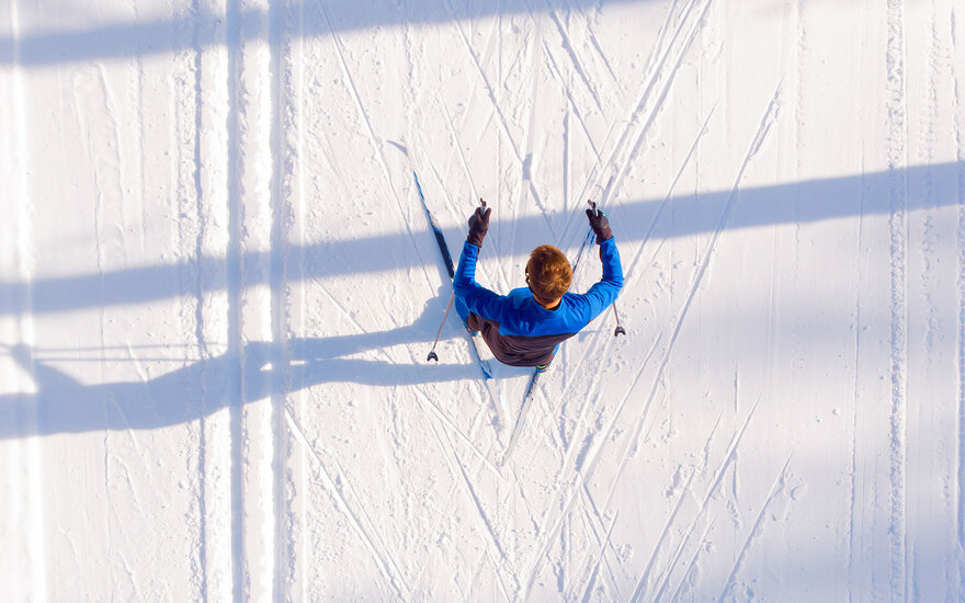 Ein Langläufer in blauer Jacke gleitet über die schneebedeckte Fläche, während Schatten von Bäumen auf den Schnee fallen.