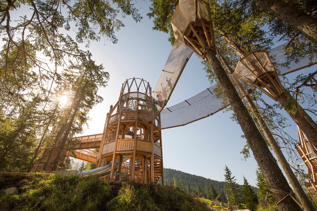 Ein beeindruckender Holzspielplatz in den Bäumen, mit Rutschen und Netzbrücken, umgeben von grünen Wäldern und Bergen.