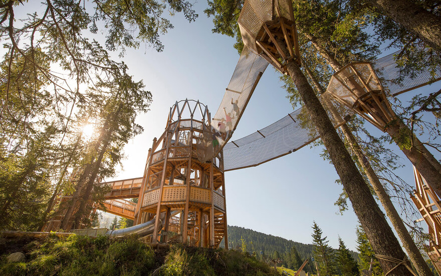 Ein beeindruckender Holzspielplatz in den Bäumen, mit Rutschen und Netzbrücken, umgeben von grünen Wäldern und Bergen.