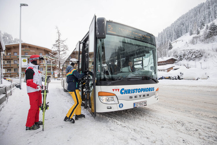 Zwei Skifahrer, einer mit Skiern, steigen in einen Bus in einer verschneiten Berglandschaft ein.