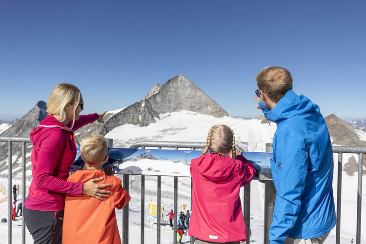 Eine Familie steht an einem Geländer und betrachtet die schneebedeckten Berge, während die Mutter auf einen Gipfel zeigt.