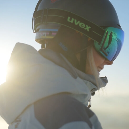 Eine Skifahrerin mit einem Helm und einer Sonnenbrille lächelt, während die Sonne hinter ihr aufgeht.
