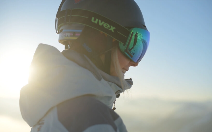 Eine Skifahrerin mit einem Helm und einer Sonnenbrille lächelt, während die Sonne hinter ihr aufgeht.