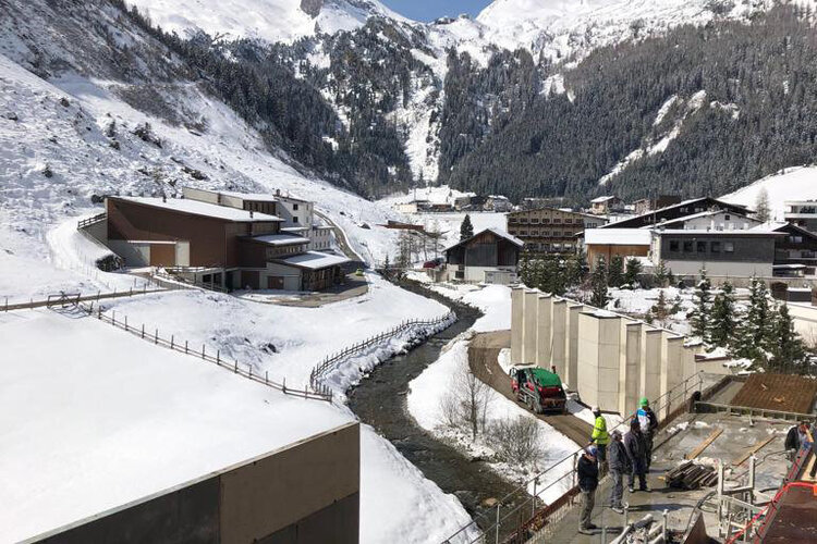 Blick auf eine schneebedeckte Berglandschaft mit einem Fluss, umgeben von modernen Gebäuden und Bauarbeitern im Vordergrund.