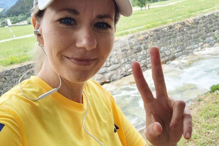Eine sportliche Frau mit Kopfhörern lächelt und zeigt ein Peace-Zeichen vor einer malerischen Landschaft.